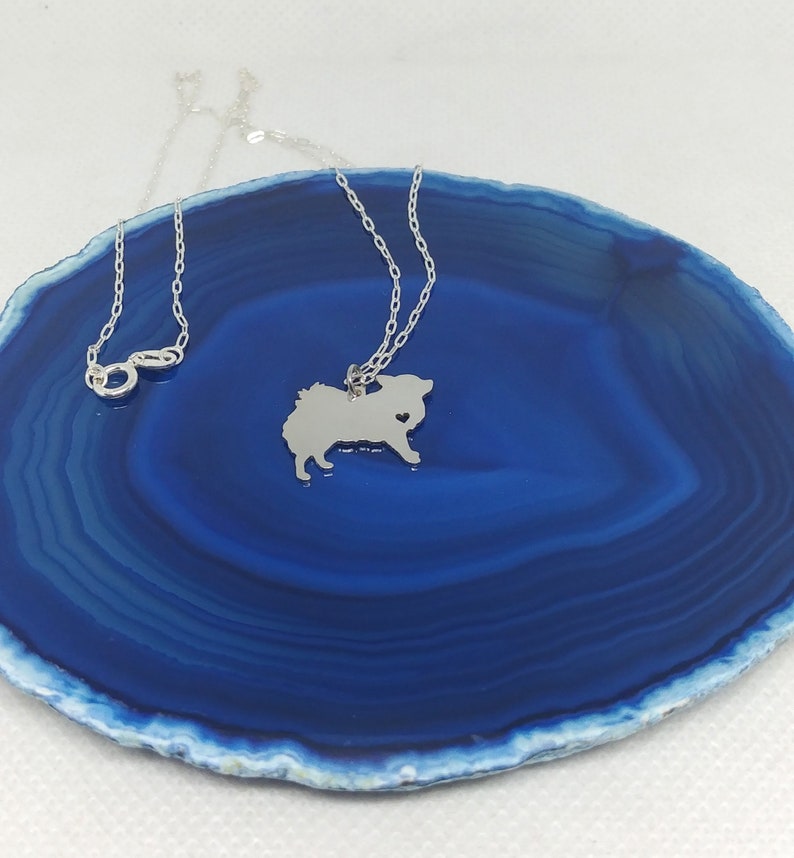 May include: A silver necklace with a small, detailed silhouette of a Pomeranian dog with a heart cut out of its chest. The necklace is on a blue agate dish.