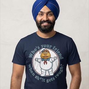 Può includere: Una t-shirt blu navy a maniche corte con la grafica di un personaggio bianco simile a un marshmallow che indossa un costume da marinaio. Il testo attorno al personaggio recita: Who's your friend when sh*!t gets rough? Design centrato sul petto.
