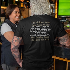 May include: A black t-shirt with a humorous saying about the golden years. The text reads: "The Golden Years Have Come At Last. I Can Not See, I Can Not Pee. I Can Not Chew, I Can Not Screw. My Memory Shrinks, My Hearing Stinks. No Sense of Smell, I Look Like Hell. My Body's Drooping. Got Troubles Pooping. The Golden Years Have Come At Last. The Golden Years Can Kiss My Ass!"