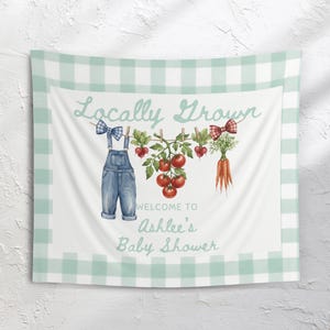 以下が含まれることがあります： 薄緑色のギンガムチェックの縁取りが付いた白いタペストリー。「Locally Grown」と「Welcome to Ashlee's Baby Shower」の文字が書かれています。デニムのオーバーオール、トマト、ニンジン、リボンなどのイラストが描かれています。