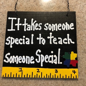 May include: Black wooden sign with white text that reads "It takes someone special to teach someone special." A colorful puzzle piece is attached to the sign. A yellow ruler is at the bottom of the sign with numbers 1 through 4.