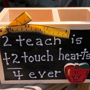 May include: Wooden box with three compartments, decorated with a yellow measuring tape and a red apple with a smiley face. The box has a chalkboard sign with the text "2 teach is +2 touch hearts 4 ever".