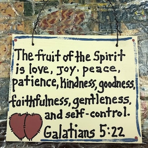 May include: A beige wooden sign with blue trim and black lettering. The sign features a heart-shaped design and the text "The fruit of the Spirit is love, Joy, peace, patience, Kindness, goodness, faithfulness, gentleness, and self-control. Galatians 5:22"