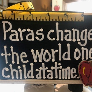 May include: A black chalkboard sign with white lettering that reads "Paras change the world one child at a time." A yellow measuring tape and a red apple with a smiley face are in the background.