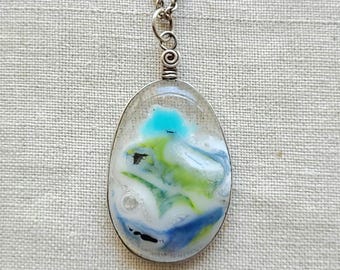 Puede incluir: Un collar con colgante de tono plateado que presenta un cabujón de vidrio ovalado con un patrón de remolinos en colores blanco, azul brillante, verde lima y azul oscuro, suspendido de una cadena de metal plateado con un engarce en espiral.