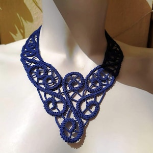Puede incluir: Un collar de encaje de ganchillo azul marino con un diseño delicado e intrincado. El collar está hecho de pequeños bucles y círculos interconectados, creando un aspecto elegante y delicado.