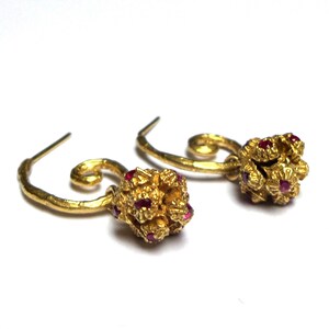 May include: A pair of gold-colored earrings with a floral design. Each earring features a cluster of small, red gemstones and a curved, hook-style closure. The earrings are set against a white background.