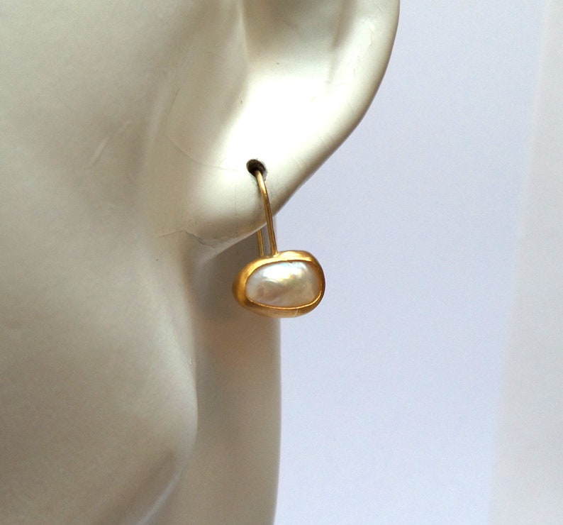 May include: A pair of gold earrings with a white pearl in a gold setting. The earrings are shown on a mannequin's ear.