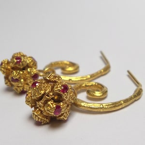May include: A pair of gold-colored earrings. Each earring features a cluster of small, round gold shapes with embedded red gemstones. The earrings have a curved, textured design and a hook closure. The background is plain white.