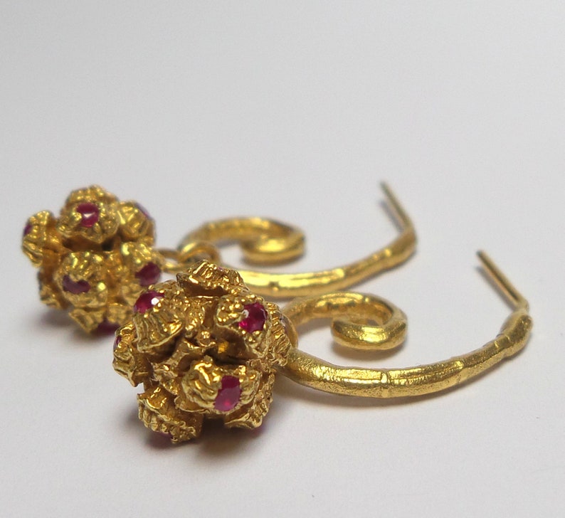 May include: A pair of gold-colored earrings. Each earring features a cluster of small, round gold shapes with embedded red gemstones. The earrings have a curved, textured design and a hook closure. The background is plain white.