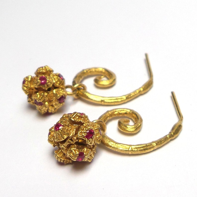 May include: A pair of gold-colored earrings with a textured, clustered design. Each earring features a small, round cluster of gold with embedded red gemstones, suspended from a curved, spiral-shaped gold wire.
