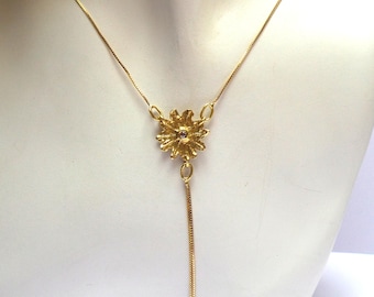 May include: A gold necklace with a delicate chain and a flower pendant with a small clear stone in the center. The pendant hangs from a long, thin chain.