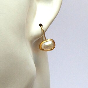 May include: Gold hoop earrings with a white pearl drop. The pearl is set in a gold bezel.