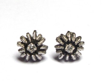 May include: A pair of silver stud earrings with a small, clear crystal in the center of each earring. The earrings are shaped like a flower with a textured design.
