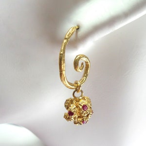 May include: A single gold-colored earring with a spiral design. The earring features a textured, irregular pendant adorned with small, red gemstones. The earring is designed to be worn in a pierced ear.