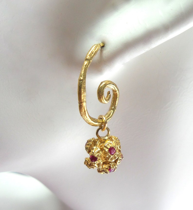 May include: A single gold-colored earring with a spiral design. The earring features a textured, irregular pendant adorned with small, red gemstones. The earring is designed to be worn in a pierced ear.
