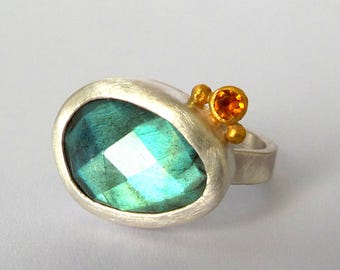 Puede incluir: Un anillo de plata con una gran piedra preciosa azul turquesa facetada engastada en un engaste de plata. El anillo tiene un pequeño acento dorado con una pequeña piedra preciosa naranja.