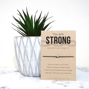 May include: A brown card with the text "You are STRONG (and don't you forget it)" and a black string bracelet with a silver star charm. The card also includes instructions for making a wish.