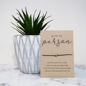 May include: A brown card with the text "you're my person" in black script. A black string bracelet with a silver star charm is attached to the card. The card is sitting on a white surface next to a potted succulent.