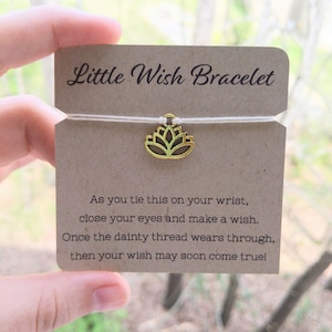 May include: A white string bracelet with a gold lotus charm. The card reads "Little Wish Bracelet" and includes instructions to tie the bracelet on your wrist, close your eyes, make a wish, and when the string wears through, your wish may come true.