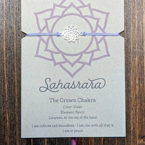 May include: A purple string bracelet with a silver charm in the shape of a flower. The card reads "Sahasrara The Crown Chakra Color: Violet Element: Spirit Location: At the top of the head I am infinite and boundless I am one with all that is I am at peace"