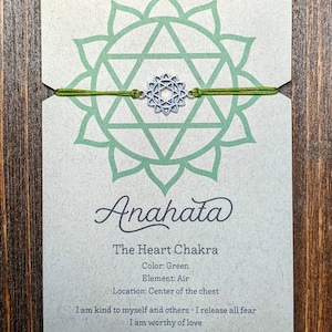 May include: Green string bracelet with a silver charm featuring the Anahata chakra symbol. The card reads "Anahata, The Heart Chakra, Color: Green, Element: Air, Location: Center of the chest, I am kind to myself and others. I release all fear. I am worthy of love."