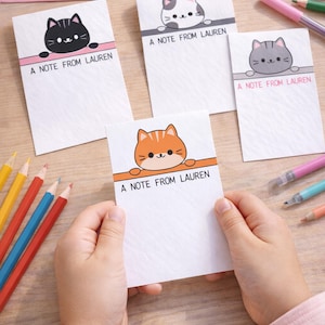 Può includere: Una collezione di biglietti bianchi con illustrazioni di gatti dei cartoni animati in nero, grigio e arancione. Ogni biglietto riporta la scritta "A NOTE FROM LAUREN". Sono visibili anche una busta rosa e matite colorate.