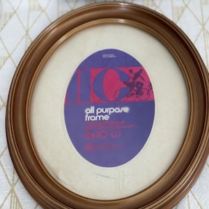 May include: An oval-shaped picture frame with a smooth, medium-brown wood finish. The frame contains a paper insert with the text "all purpose frame" and "8x10 5x7" printed in white and red against a purple background with a bicycle graphic.