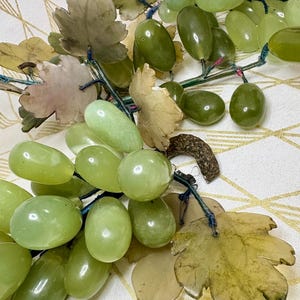 May include: A cluster of green, oval-shaped grapes with detailed leaves. The grapes vary in shades of green, and the leaves are a mix of green and tan. The stems are a dark blue color. The grapes are arranged on a white and gold patterned surface.