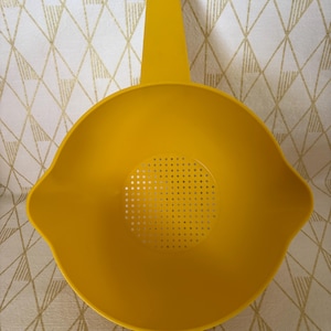 May include: A bright yellow plastic colander with a handle. The colander has a round bowl shape with a perforated bottom and a long handle for easy use. The handle has a hole for hanging. The colander is designed for draining liquids from food.