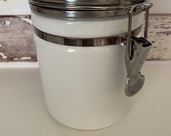May include: White ceramic canister with a silver lid and a silver latch. The canister has a silver band around the middle.