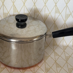 May include: A stainless steel saucepan with a copper-colored base, featuring a matching lid with a black knob and a long black handle with a small metal hanging loop at the end. The pot has a polished metallic finish and a cylindrical shape.
