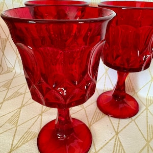 May include: Four vintage red glass goblets. The goblets have a diamond pattern on the bowl and a round base. The stemware is a vibrant red color, and the glasses are arranged on a patterned surface.