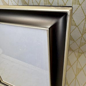 May include: A corner view of a rectangular picture frame. The frame has a black interior and a silver-colored exterior. The frame is empty, with a white interior. The background is a cream color with a gold geometric pattern.