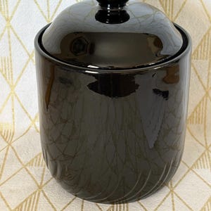 May include: A glossy black ceramic canister with a matching lid featuring a rounded knob handle. The cylindrical container has a smooth finish on the upper portion and subtle vertical ribbed detailing along the bottom edge of the exterior.