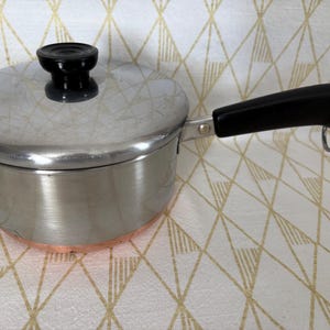 May include: A small, stainless steel saucepan with a lid and a black handle. The pot has a copper-colored bottom and a black knob on the lid. The handle has a metal loop at the end.