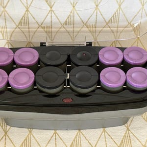 May include: An electric hair roller set featuring a black base with a clear hinged lid. The unit contains twelve cylindrical rollers arranged in two rows, with eight purple rollers on the outer edges and four black rollers positioned in the center.