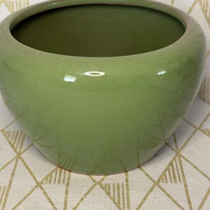 May include: A round, light green ceramic planter with a glossy finish. The planter has a wide opening and a slightly flared rim. The interior is a darker shade of green. The planter is likely for indoor or outdoor use.