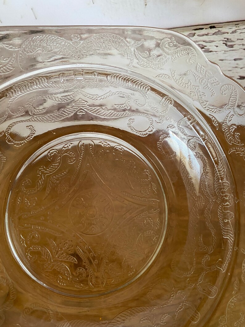 Peut inclure: Une assiette en verre transparent avec un bord festonn&eacute; et un motif grav&eacute; complexe de feuilles et de vignes tourbillonnantes. L'assiette a une teinte l&eacute;g&egrave;rement ambr&eacute;e.