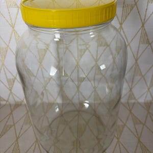 May include: A large, clear glass jar with a bright yellow lid. The jar has a wide body and a narrow neck, suitable for storage. The lid is made of plastic and screws on tightly. The jar is empty and ready to be filled.