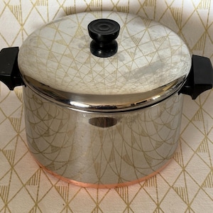 May include: A stainless steel stockpot with a polished finish and a copper-colored base. The pot features two black side handles and a matching lid with a black circular knob on top. The metal surface reflects the surrounding geometric pattern.