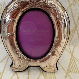 May include: An oval silver-plated picture frame featuring embossed floral and vine detailing along the border. The center contains a purple insert with the text Ashton & Hayes and Silver Plated printed in a light script font.