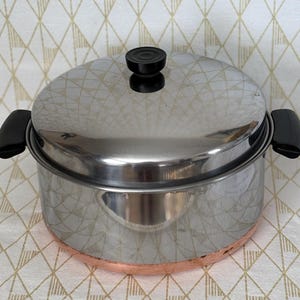 May include: A stainless steel cooking pot with a matching lid, featuring black plastic handles on the sides and a black knob on the lid. The base of the pot is made of copper, and the polished metal surface reflects the surrounding environment.