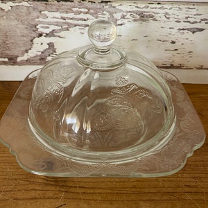 Peut inclure: Un beurrier en verre transparent avec un couvercle en forme de d&ocirc;me et un motif floral. Le plat a un bord festonn&eacute; et est pos&eacute; sur une surface en bois.