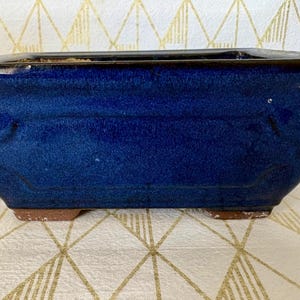 May include: A rectangular ceramic bonsai pot with a deep cobalt blue glazed finish. The pot features a flared rim, slightly tapered sides with subtle embossed panel detailing, and four small unglazed terracotta feet at the base.