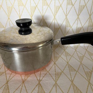 May include: A stainless steel saucepan with a lid and a black handle. The pot has a copper-colored bottom and a black knob on the lid. The handle has a metal loop at the end. Kitchenware.
