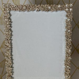 May include: A rectangular picture frame featuring a metallic silver border densely encrusted with numerous clear, round, faceted rhinestones of varying sizes. The frame has a plain, light-colored, rectangular center insert for displaying a photograph.