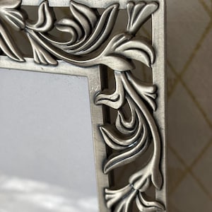 May include: A rectangular picture frame with a metallic silver finish. The frame features an intricate, raised relief pattern of scrolling acanthus leaves along the outer border, surrounding a plain, light-colored rectangular center insert.