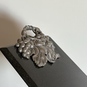 May include: A black rectangular plaque with a silver-colored grape and leaf design. The decorative element is attached to the top of the plaque. The plaque is angled, revealing a glimpse of its polished surface.