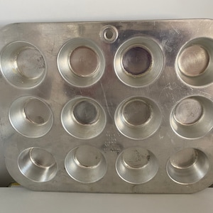 May include: A silver metal muffin tin with 12 individual muffin cups. The tin is slightly worn and has a few scratches.
